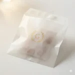 Close-up of elegant glassine paper bag surface showing natural paper gloss, gold brand monogram print, and soft product silhouette through semi-translucent paper