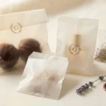 Elegant glassine packaging bags in boutique retail scene with dried flower and wax seal props — custom-printed semi-translucent paper bags for artisan food, cosmetics, and gift brands